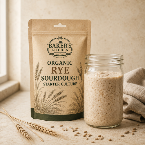 A kraft paper pouch labeled 'The Baker's Kitchen Organic Rye Sourdough Starter Culture' stands next to a glass jar filled with a beige sourdough starter culture, styled with wheat stalks and seeds on a warm stone surface.